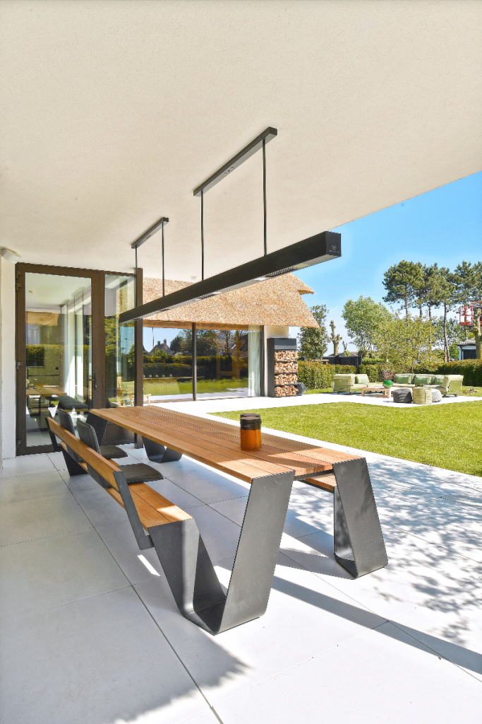 an outdoor kitchen design featuring a warm oak and grey metal bench and table on concrete paving below a long grey pendant light in a natural green setting