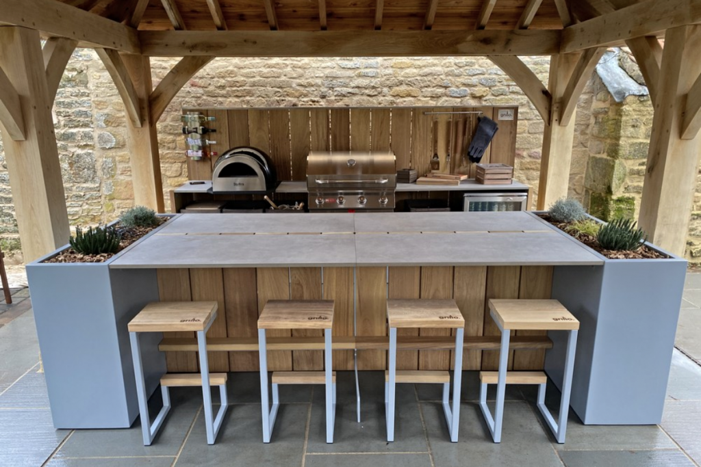 an outdoor kitchen design featuring exposed brickwork, tables and planters painted in a blue-grey shade and wooden bar stools