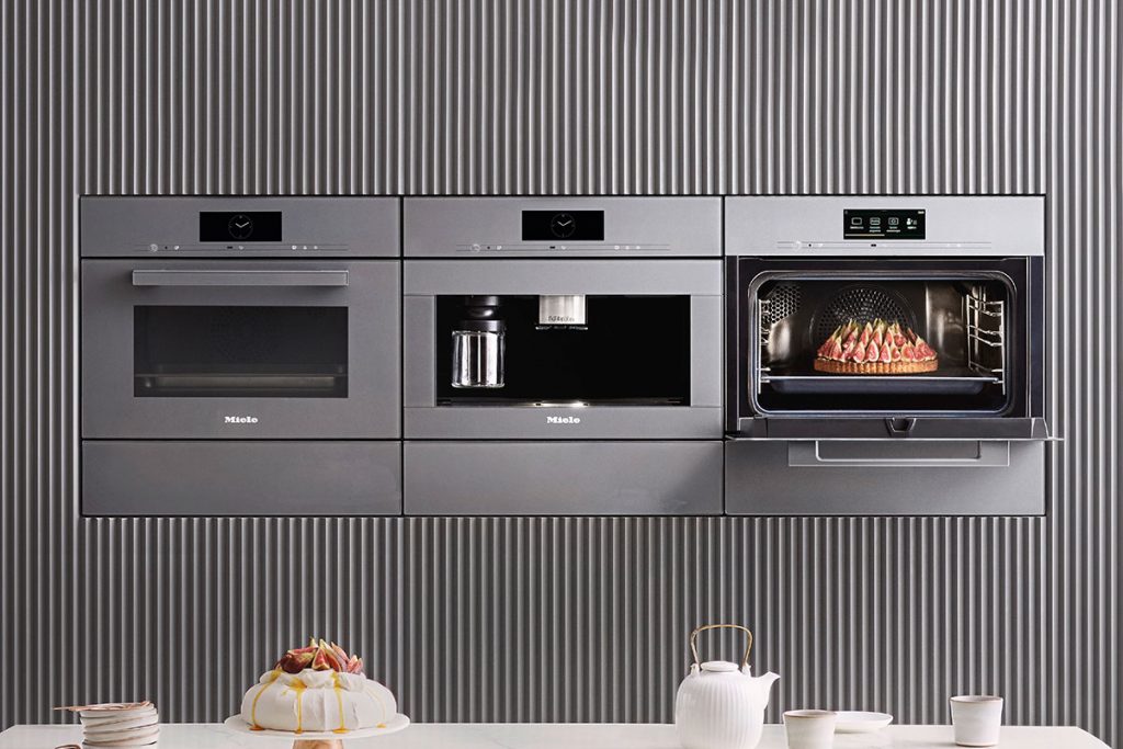 a row of three stainless steel Miele ovens, two closed and one with a Pavlova dessert on display, in front of an island set for a party, to illustrate a feature on kitchen must-haves for entertaining