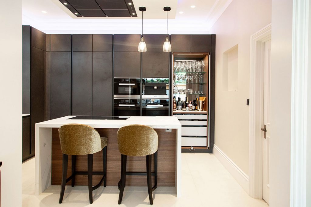 gold bar stools behind a white and walnut kitchen island in front of a dark wood home bar to illustrate a feature on kitchen must-haves for entertaining
