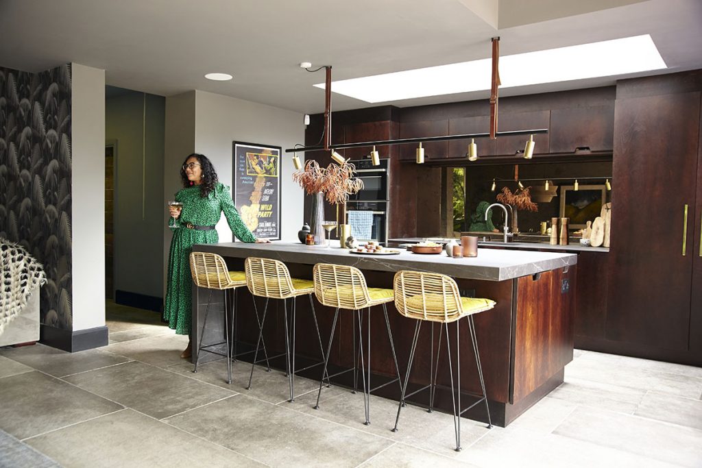 a woman in a green dress standing holding a cocktail glass next to yellow bar stools at a grey and walnut wood island with brass pendant lights above it