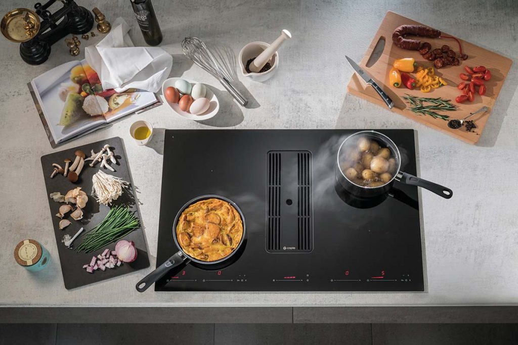 a black induction hob with a red display and two pans on it cooking potatoes and a quiche, with lots of cooking accessories and food around it