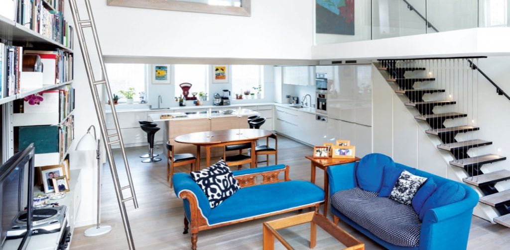 a two-level space with an illuminated stairway up to the second level, featuring blue sofas, several bookshelves and a white island with black bar stools around it