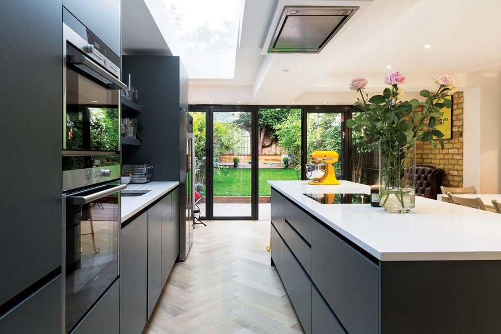 monochrome cabinetry and fittings in an island in front of bifold doors, with a bunch of pink roses in a vase and a yellow electric mixer on top of the island
