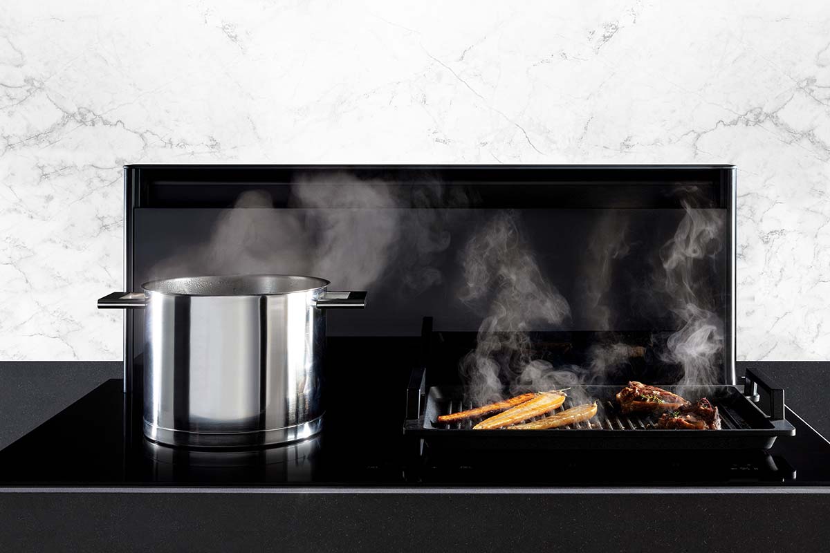 a black downdraft extractor with a steaming pan and steaming fish and meat on a grill pan 