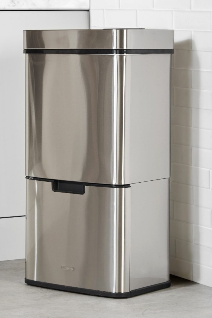 a silver freestanding three-bin disposal unit with a black rim standing on its own against a white brick wall