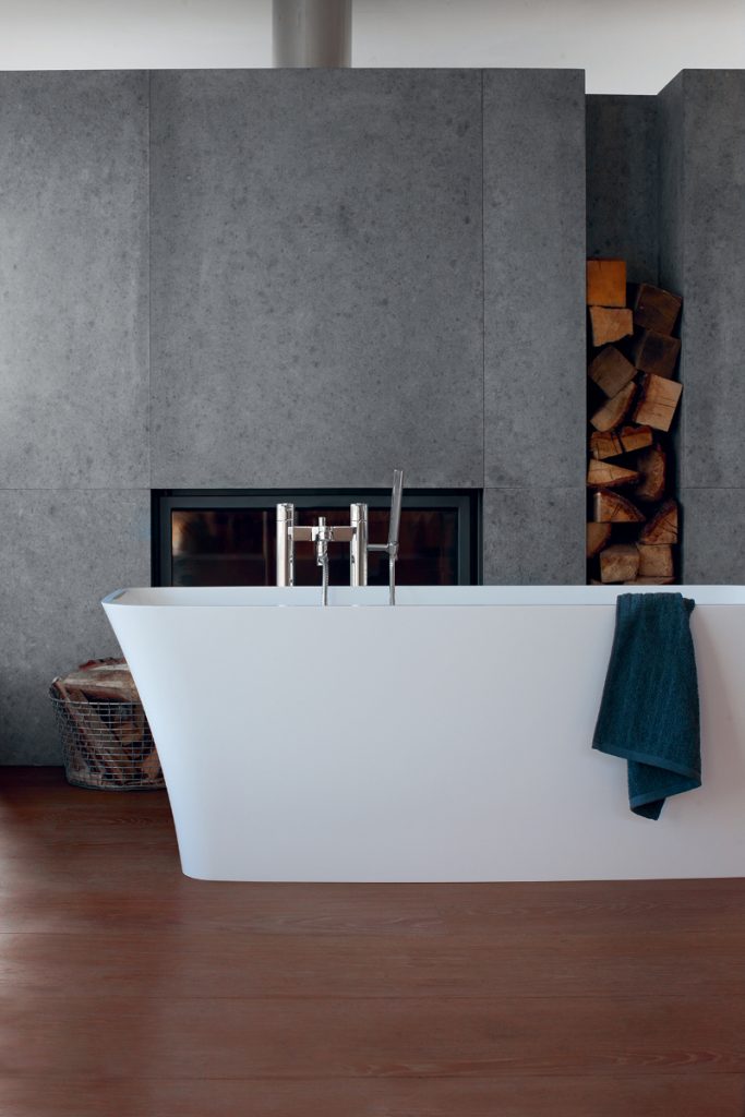 a modern rustic bathroom with a freestanding bath with silver taps against a grey stone wall and fireplace