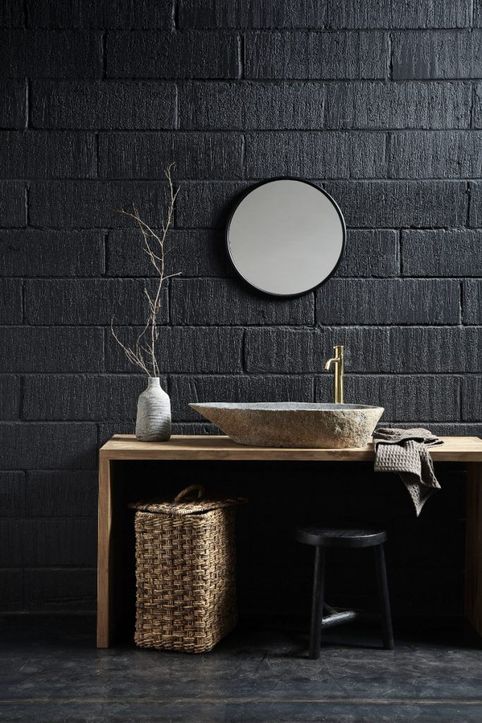 a round stone basin on a wooden countertop with a brass tap against a black brick wall