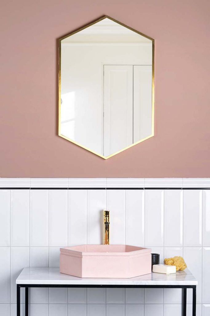 a maximalist bathroom featuring a hexagonal pastel pink basin on a marble counter top with a brass tap and brass mirror above it