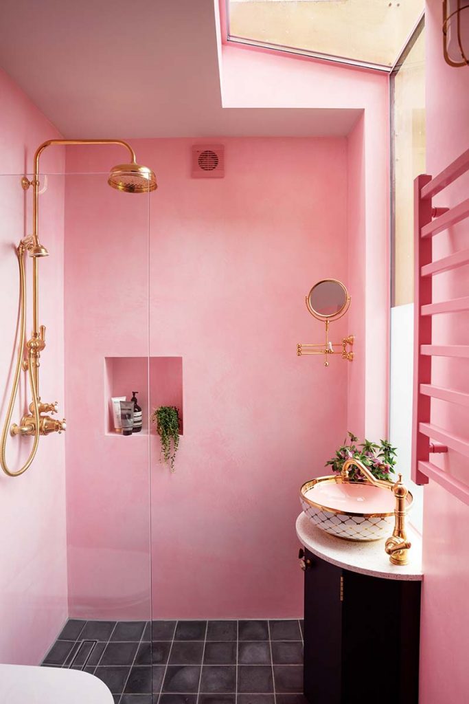 a baby pink maximalist bathroom with a brass shower, a pink basin and a black tiled floor