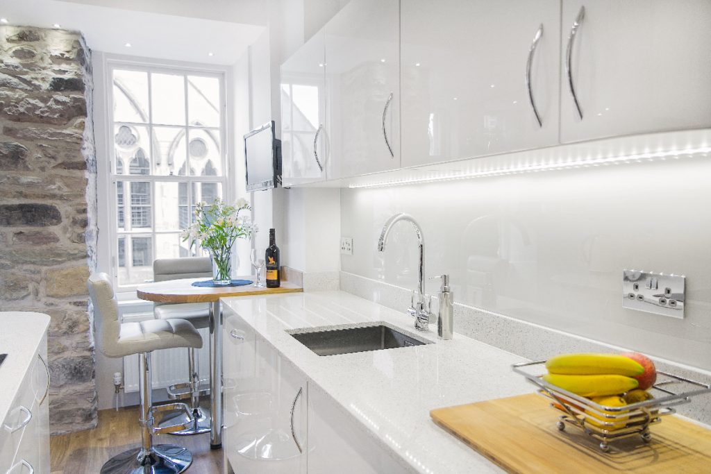 a small kitchen painted white with silver handles next to an oak breakfast bar with two bar stools