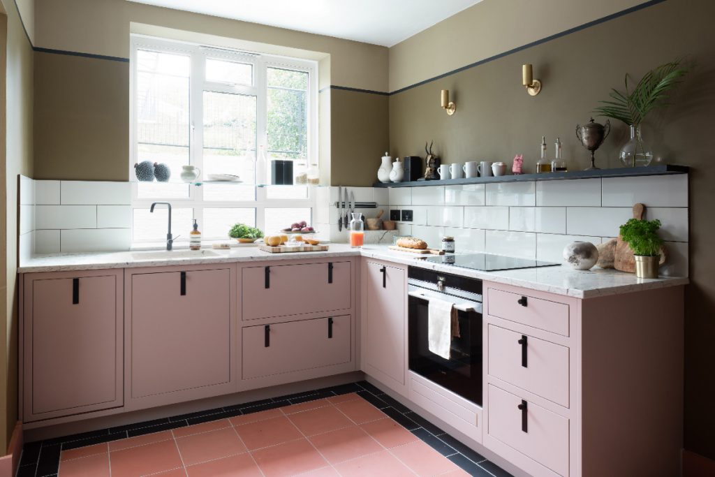 a small pink kitchen with black pull handles and white metro brick tiling with open shelving above