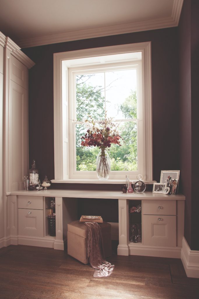 a built in table for getting ready at with perfume bottles and pictures on top and a big vase of flowers by a window