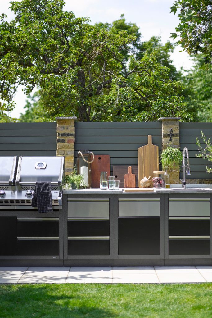 painted grey fences above stainless steel cabinetry and appliances accessorised with wooden chopping boards, a sink and a stainless steel tap