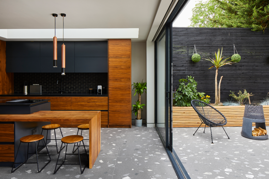an outdoor kitchen design made of stained wood with pendant lights, a wooden kitchen island with wooden bar stools underneath, bifold doors and terrazzo floor tiles 