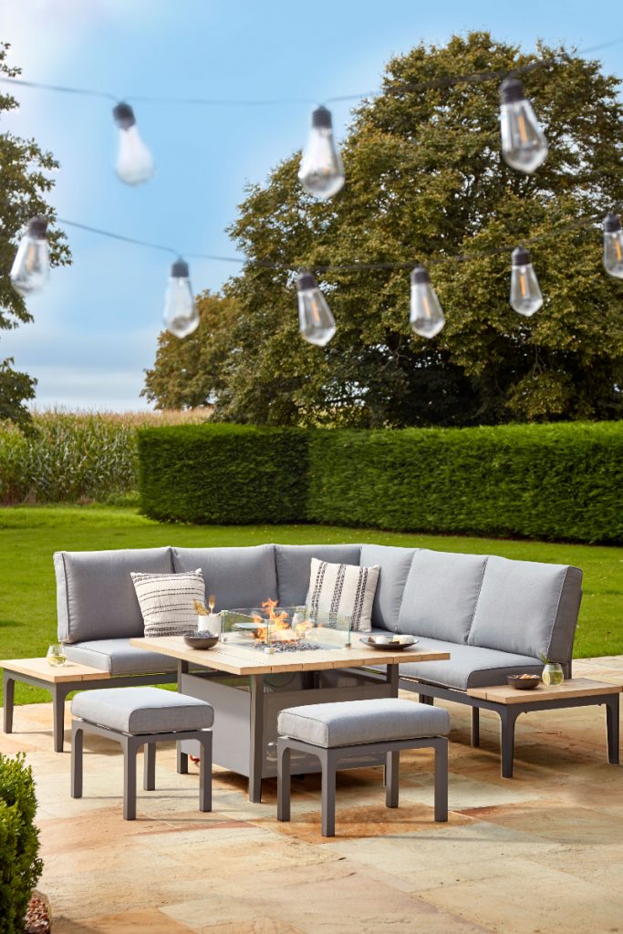 the Stockholm corner lounge sofa with grey cushions and seating and a table in the middle featuring a fire pit, against backdrop of green hedges