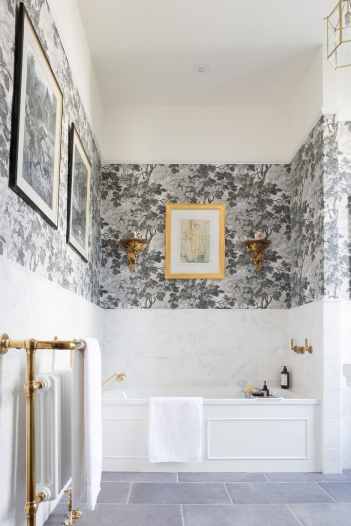a traditional bathroom with a white fitted panelled bath, floral wallpaper and several traditional pictures