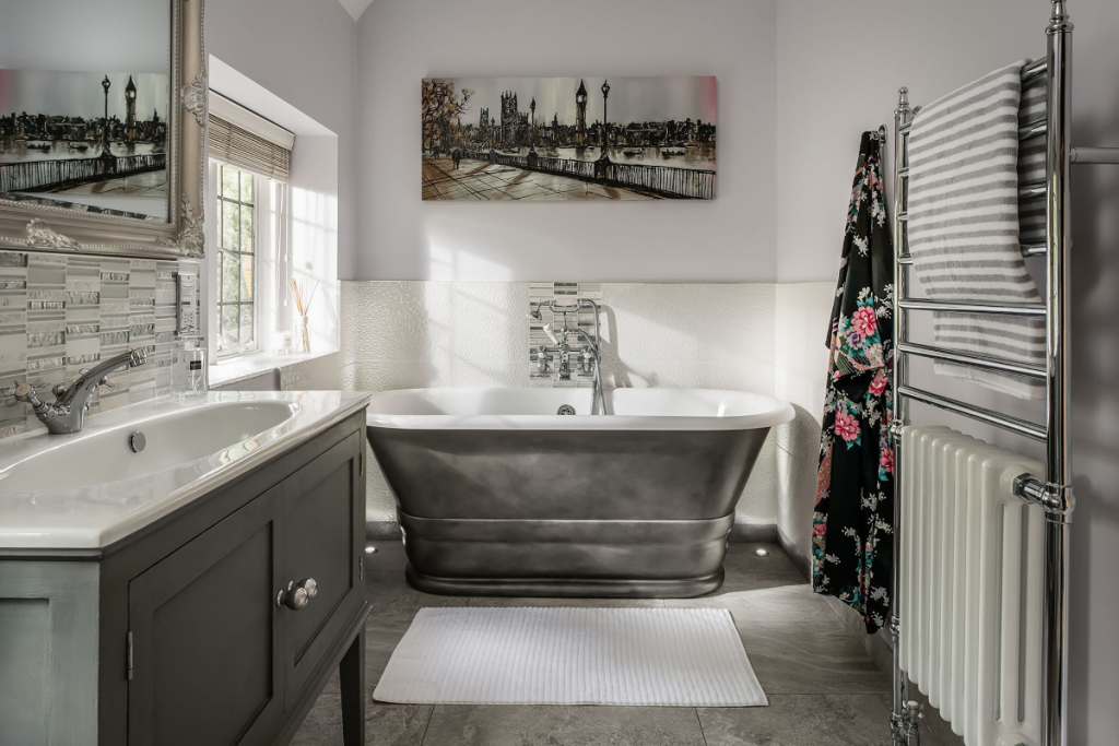 a metallic roll top bath with a silver shower, a large picture of London, a silver towel rail and a pedestal basin