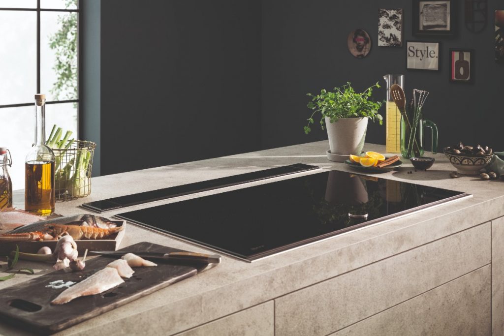 the Neff TwistPad Fire control cooktop built into a beige stone kitchen island, with assorted accessories around it including plants, utensils, food and chopping boards