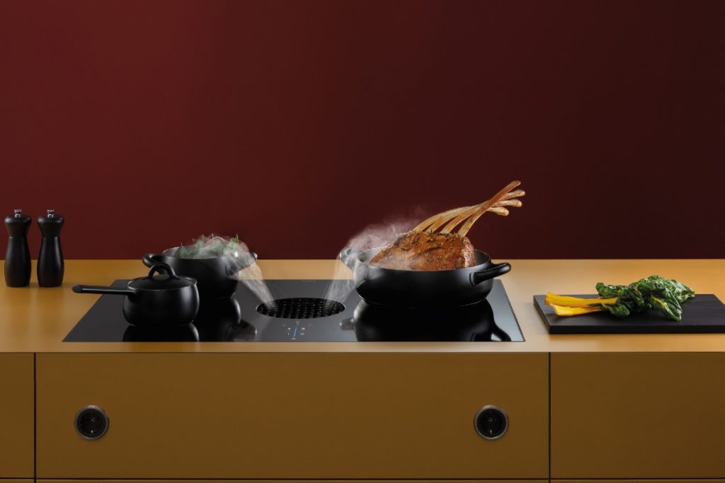 a hob with extractor in yellow worktop in a kitchen with two black pans containing chicken and vegetables steaming on top of it. There is a chopping board featuring pak choi to the right of it and two salt and pepper shakers to the left