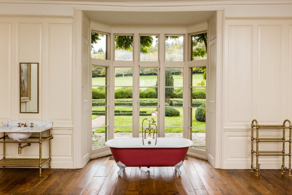 a heated brass towel rail next to a red roll top bath in front of a large window