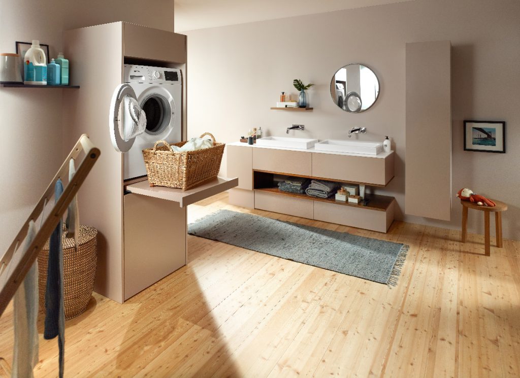 a laundry room with a wooden plank floor, twin sinks, chrome taps, a round mirror, taupe cabinetry, a washing machine and two rattan baskets