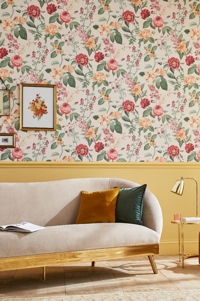 the latest wallpaper from Graham & Brown featuring flowers in red, orange and green shades, behind a brass sofa with green and orange cushions