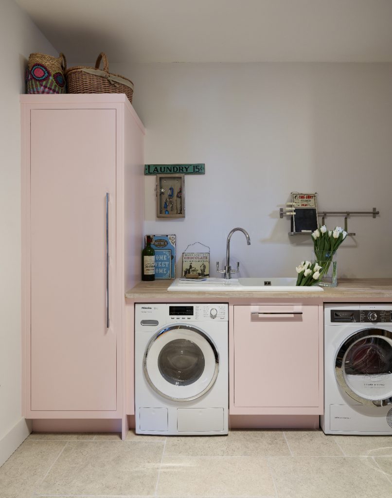 a laundry room with pastel pink cabinetry, silver handles, a washing machine, a tumble dryer, and two bunches of white tulips in a white undercounted sink with chrome tap