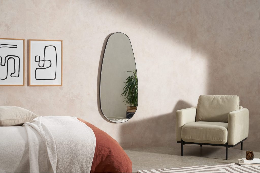a large mirror in a neutral room with a beige armchair and black and white pictures on the wall