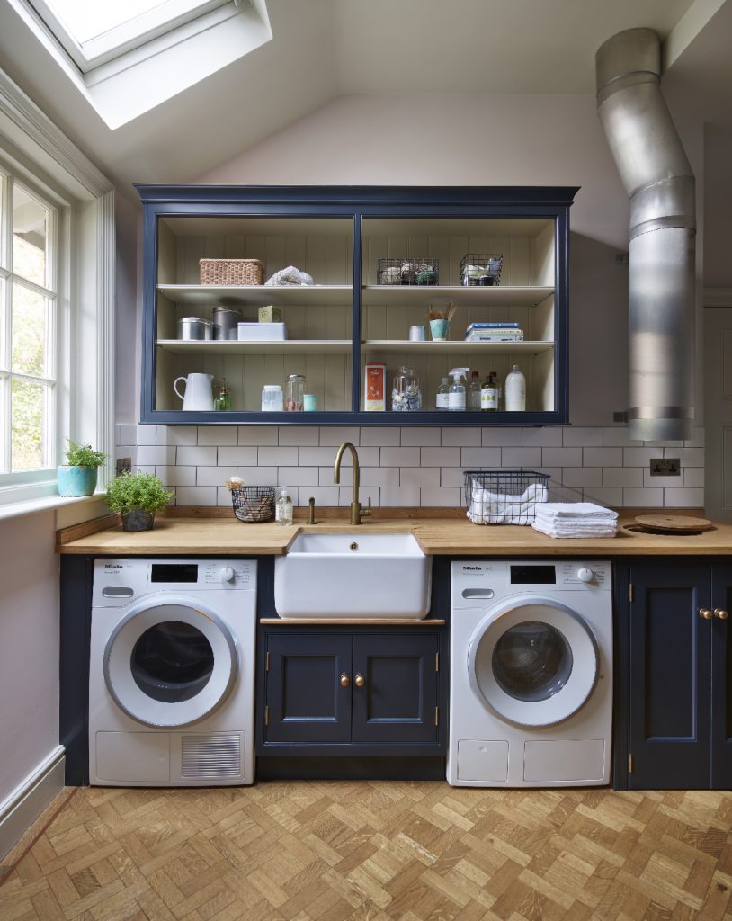 twin washer dryers, a white Belfast sink with brass tap, navy cabinetry, a wooden worktop and white metro tiles