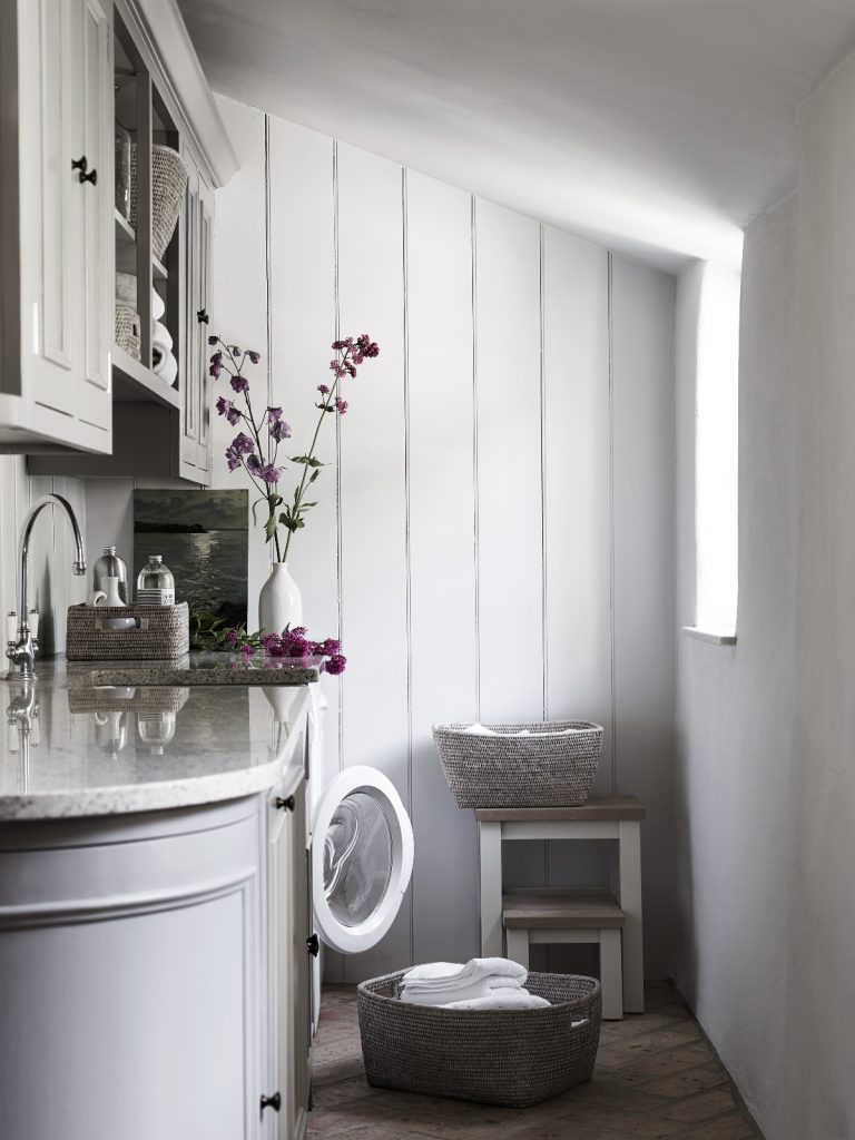 a white laundry room featuring purple flowers, a washing machine with an open door, several wicker baskets, a sink with silver tap and a window