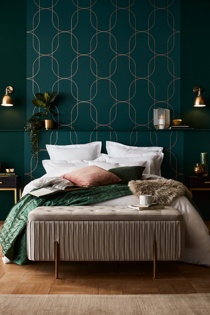 the latest wallpaper from Graham & Brown in a green and gold geometrical design behind a double bed with pink and green bedding and a beige ottoman