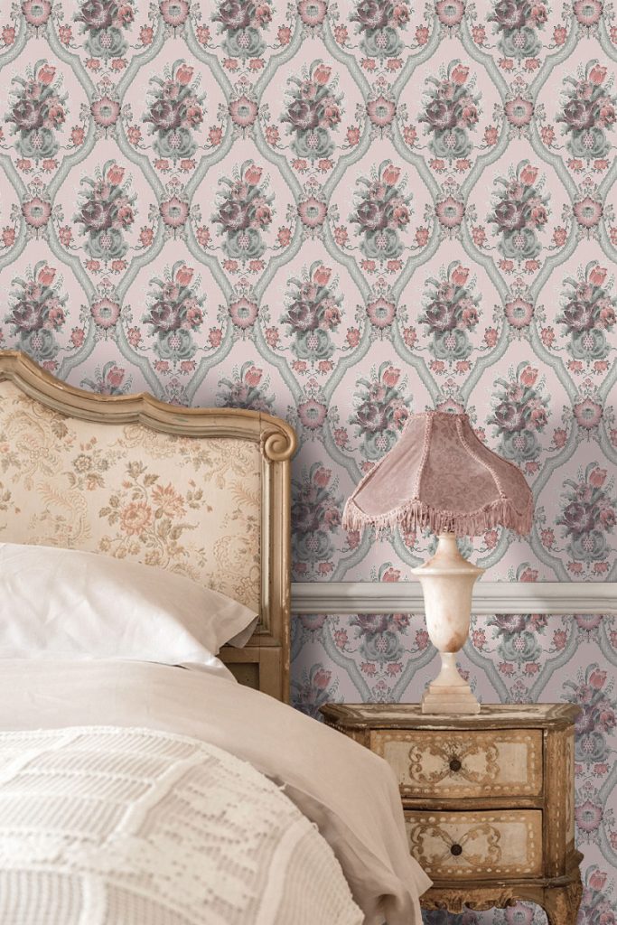 a floral print in pink and green behind an antique double bed with a gold nightstand and pink lamp