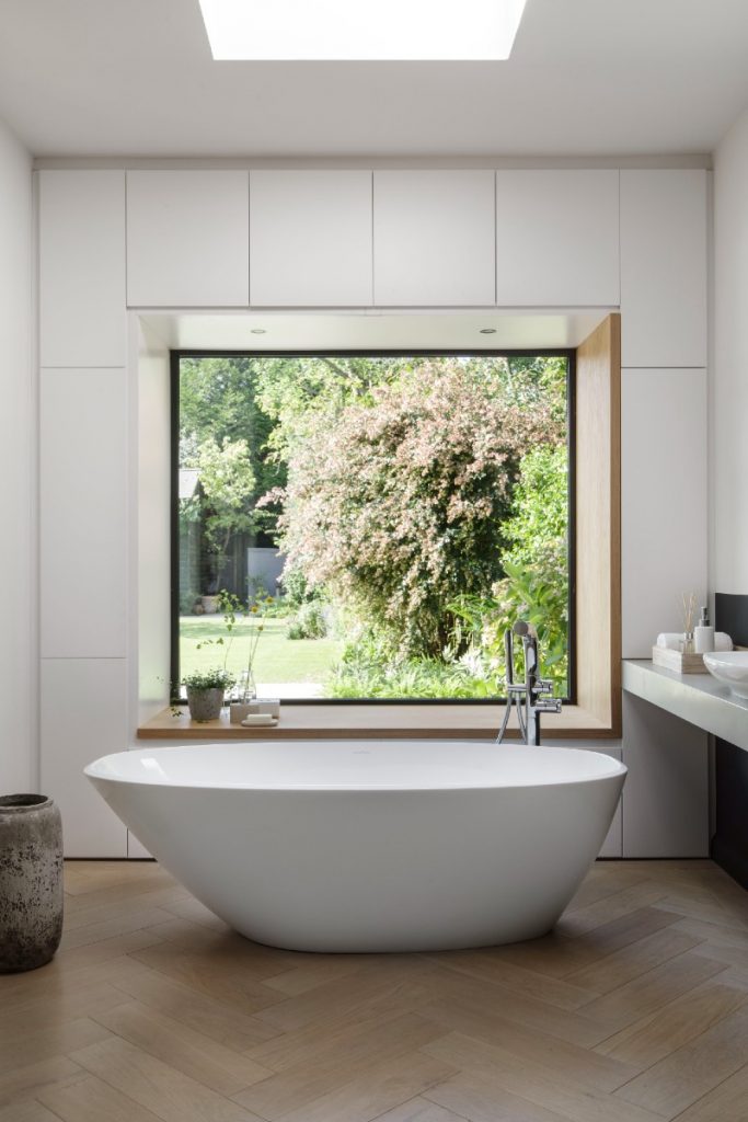 a white oval double‐ended bath in front of a square picture window in a bathroom featuring herringbone flooring 