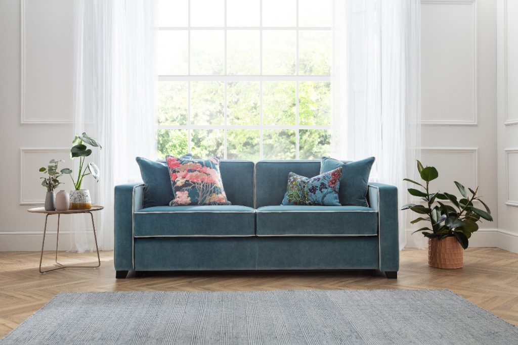 a teal double sofa bed with floral cushions in a spacious living room with wooden herringbone flooring, plants and a grey rug