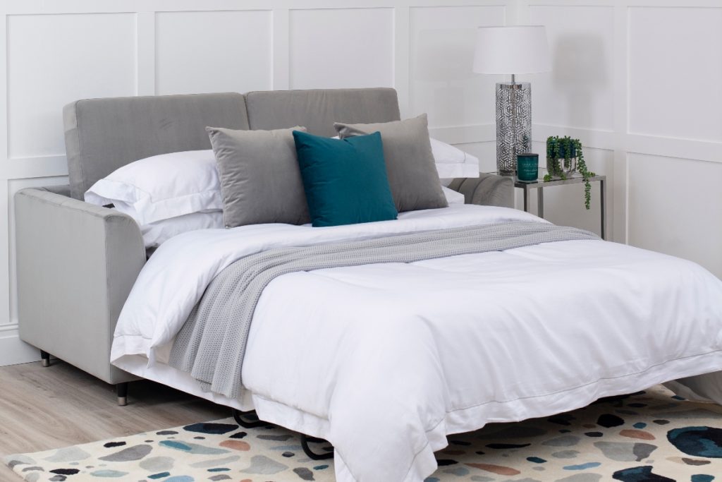 a grey double sofa bed with grey and teal cushions next to a grey and white table lamp and a trailing plant