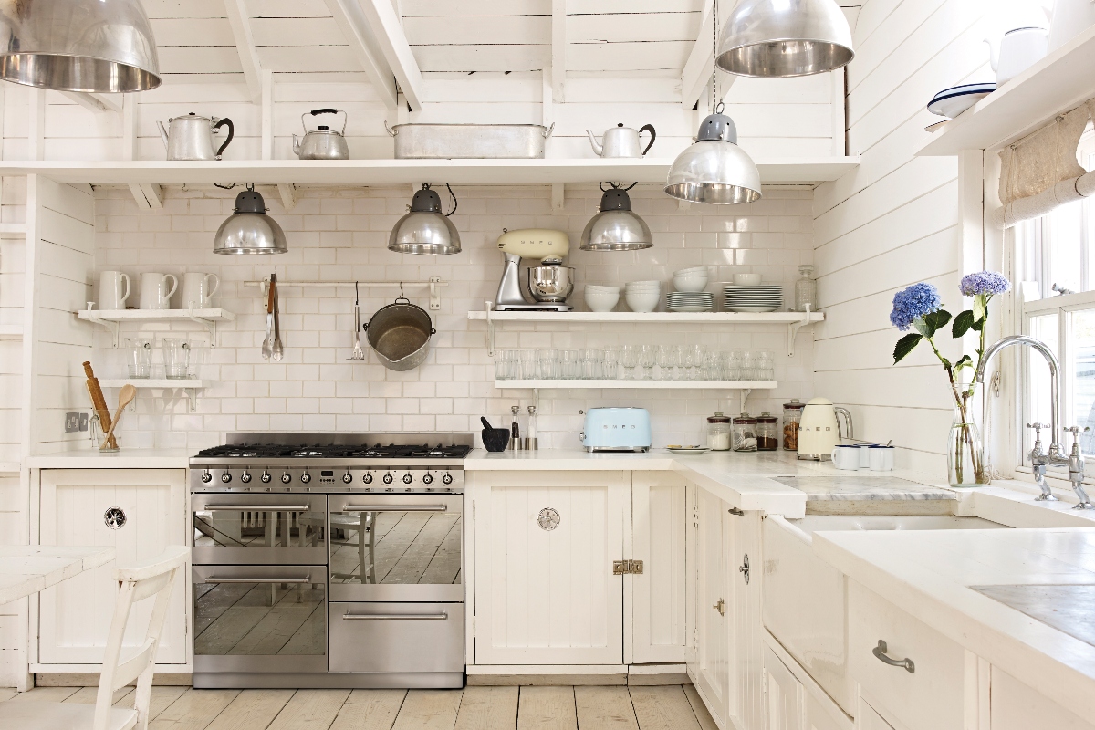 a country-style design with open shelving, cream cabinetry, a pastel blue Smeg toaster, lots of drinking glasses displayed and a Belfast sink with a silver tap