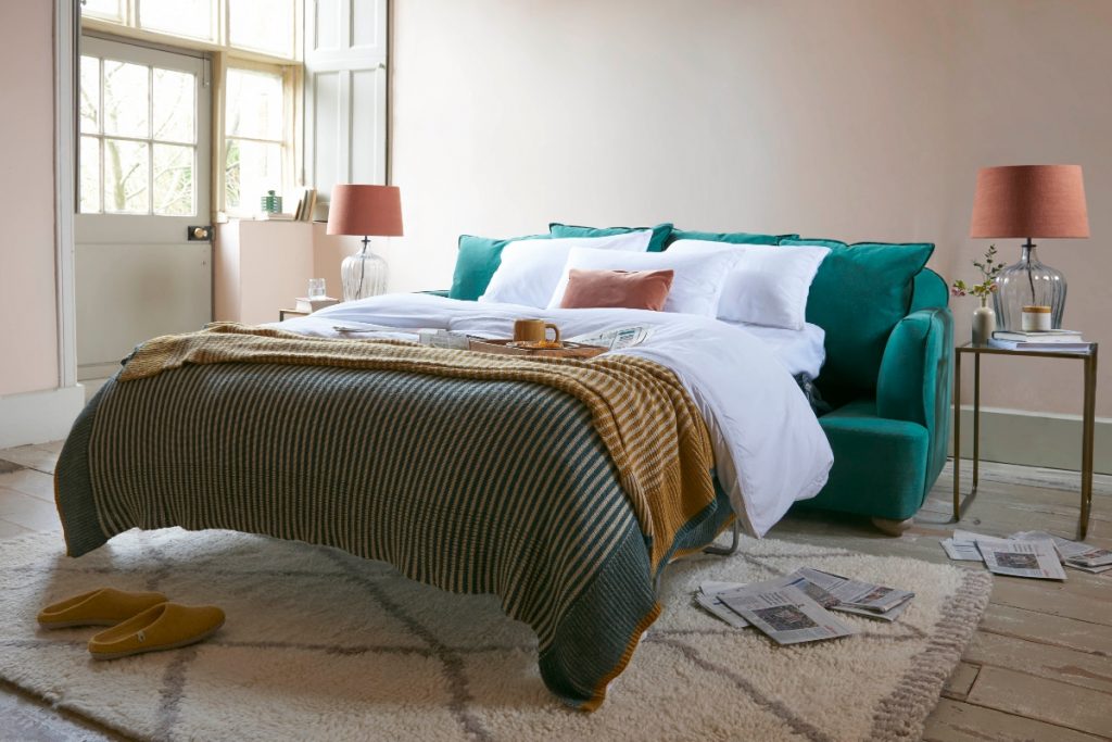 a teal double sofa bed with green and white bedding on a beige rug and two adjacent table lamps with orange shades