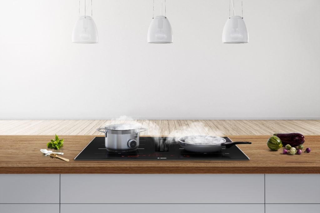 a black induction hob with built-in extractor with two pans steaming on it and three pendant lights above it