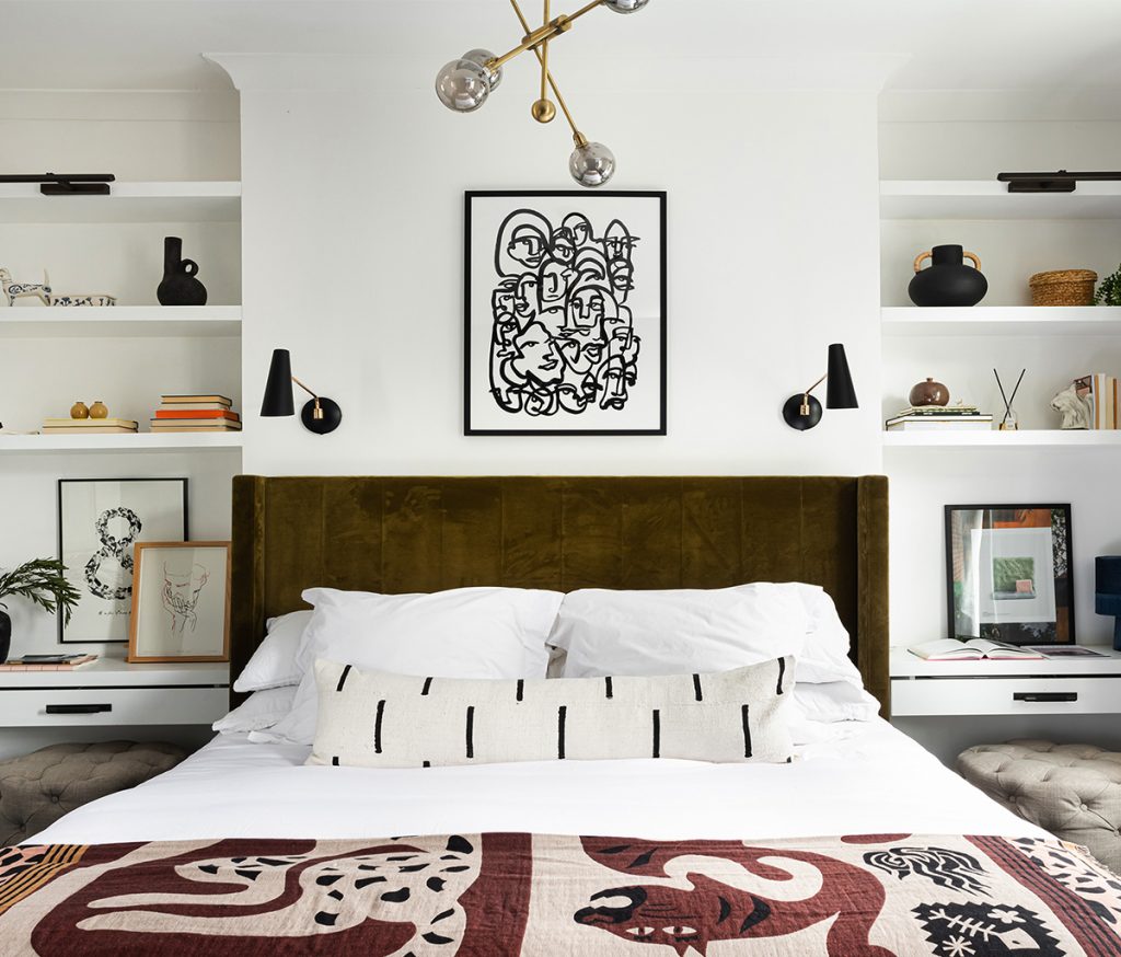 a bedroom featuring a brown velvet double bed, a patterned throw and picture, two black wall lamps and a selection of accessories on white shelves
