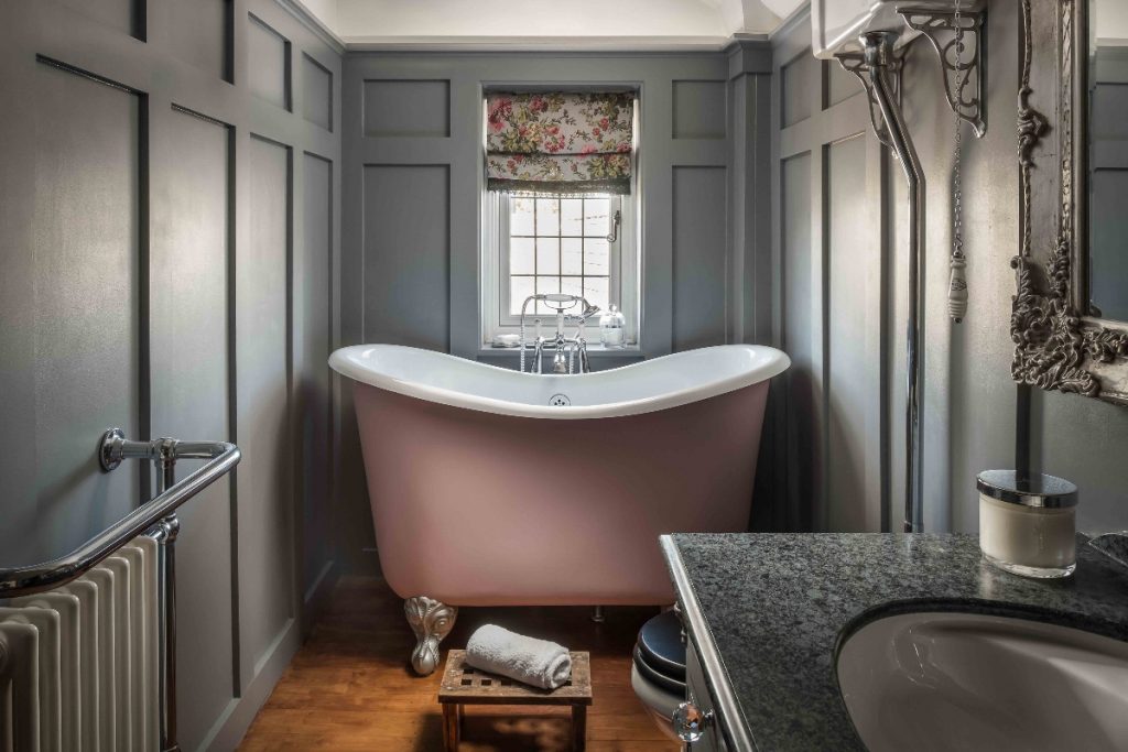 a freestanding taupe compact bath in a grey panelled bathroom with toiletries and accessories 