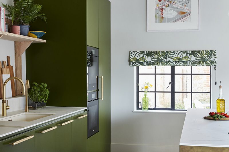 army green cabinetry with brass handles and a steel framed window with a tropical print blind