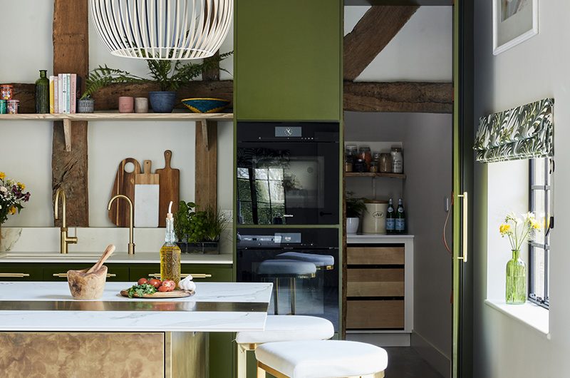 a Sola Kitchens design featuring wooden beams, army green cabinetry and brass bar stools around a brass and marble kitchen island