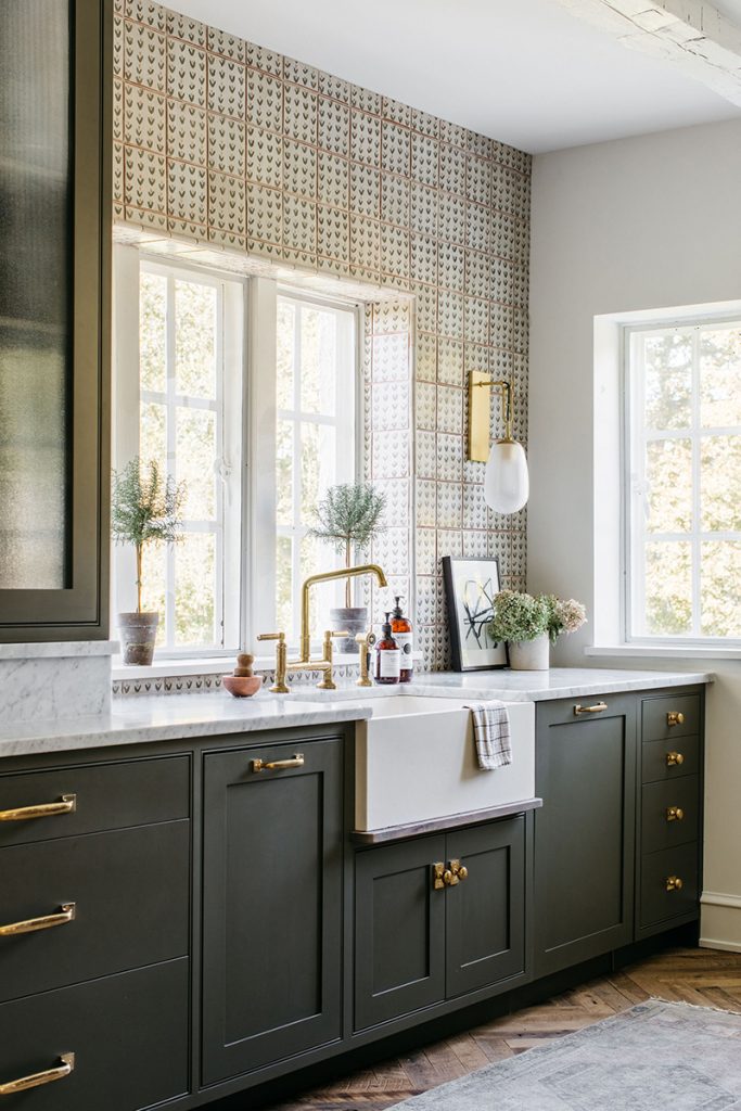 a kitchen with olive green shaker cabinetry with brass handles, a white Belfast sink and brass tap, and floral tiles