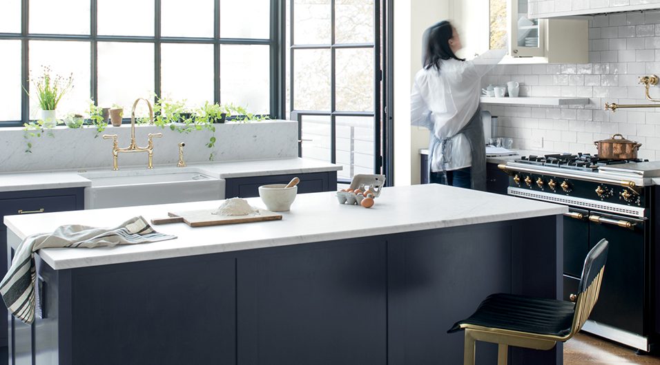 a kitchen with navy handleless cabinetry, a marble island with brass bar stool, steel framed glazing and a woman getting crockery from a cupboard, to illustrate the question of how much a kitchen costs