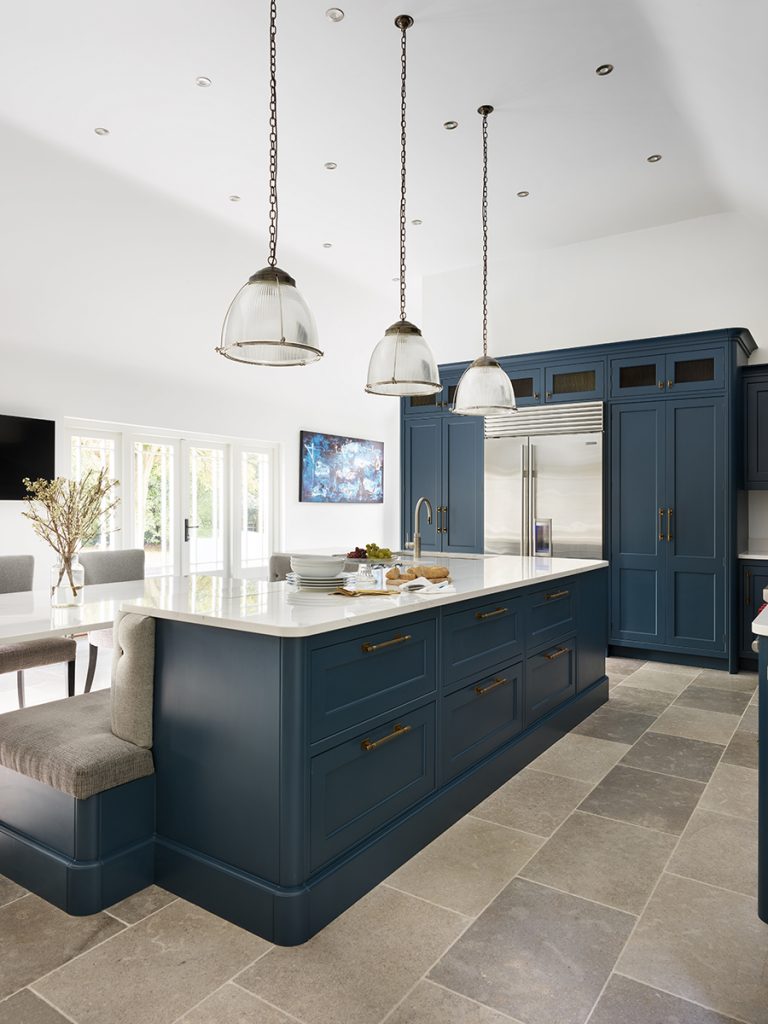 a navy kitchen with banquette seating and brass handles with three pendant lights over a white marble island