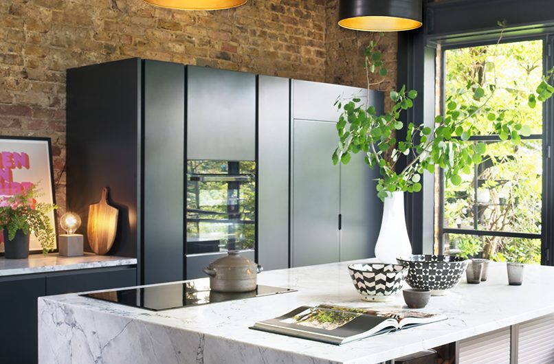 a kitchen with black pendant lights above a white marble island with appliances in navy cabinetry in the background against exposed brickwork