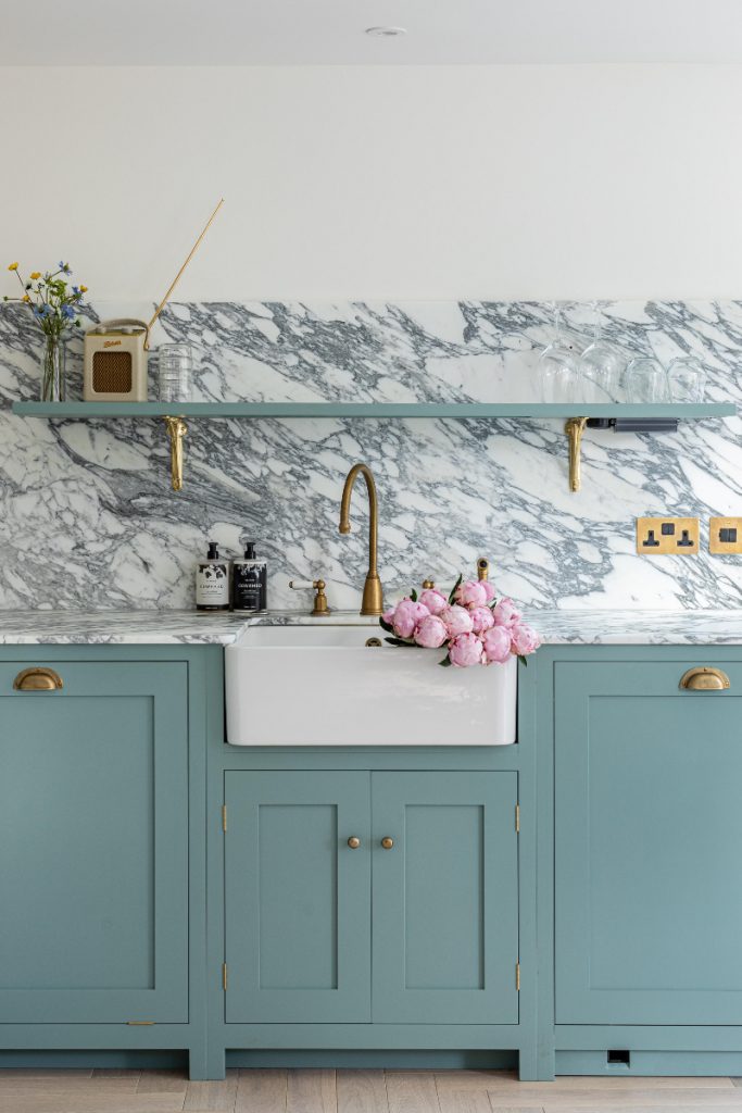 a Belfast sink with a brass tap and a bouquet of pink roses in it against a veined marble splashback above teal Shaker cabinetry with brass handles