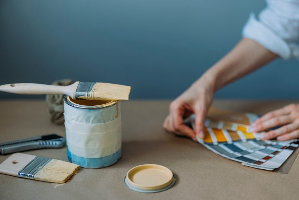 a woman holding paint swatches next to an open paint pot as she's painting her own house due to the pingdemic