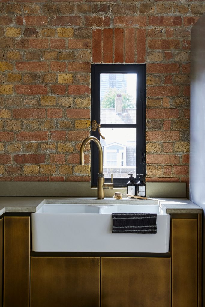 a Belfast sink set into brass cabinetry with a brass tap in front of exposed brickwork
